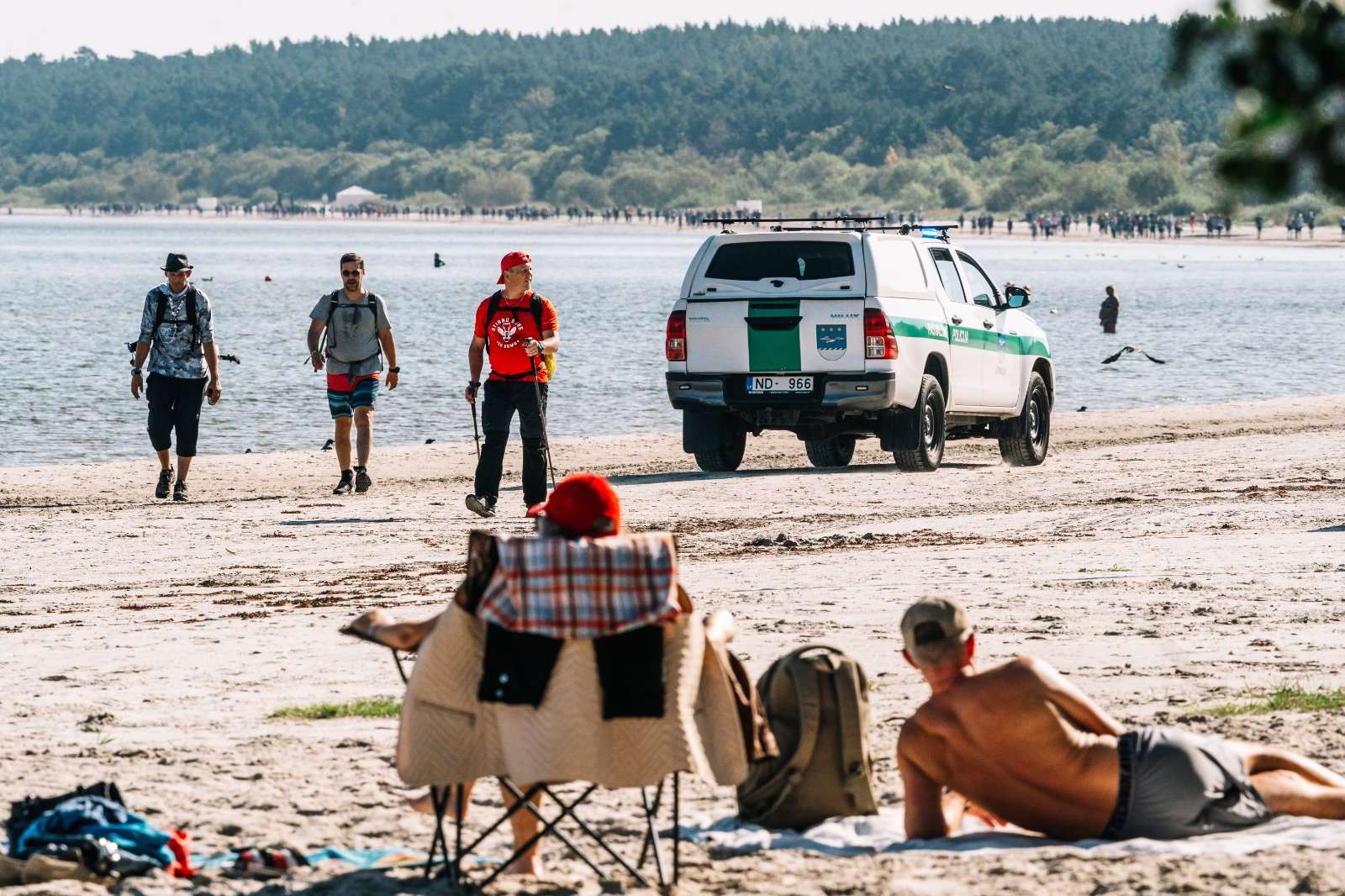 Trīs pārgājiena dalībnieki pludmalē soļo Kolkas virzienā, priekšplānā zviln atpūtnieki, fonā Jūrmalas pašvaldības policijas auto.